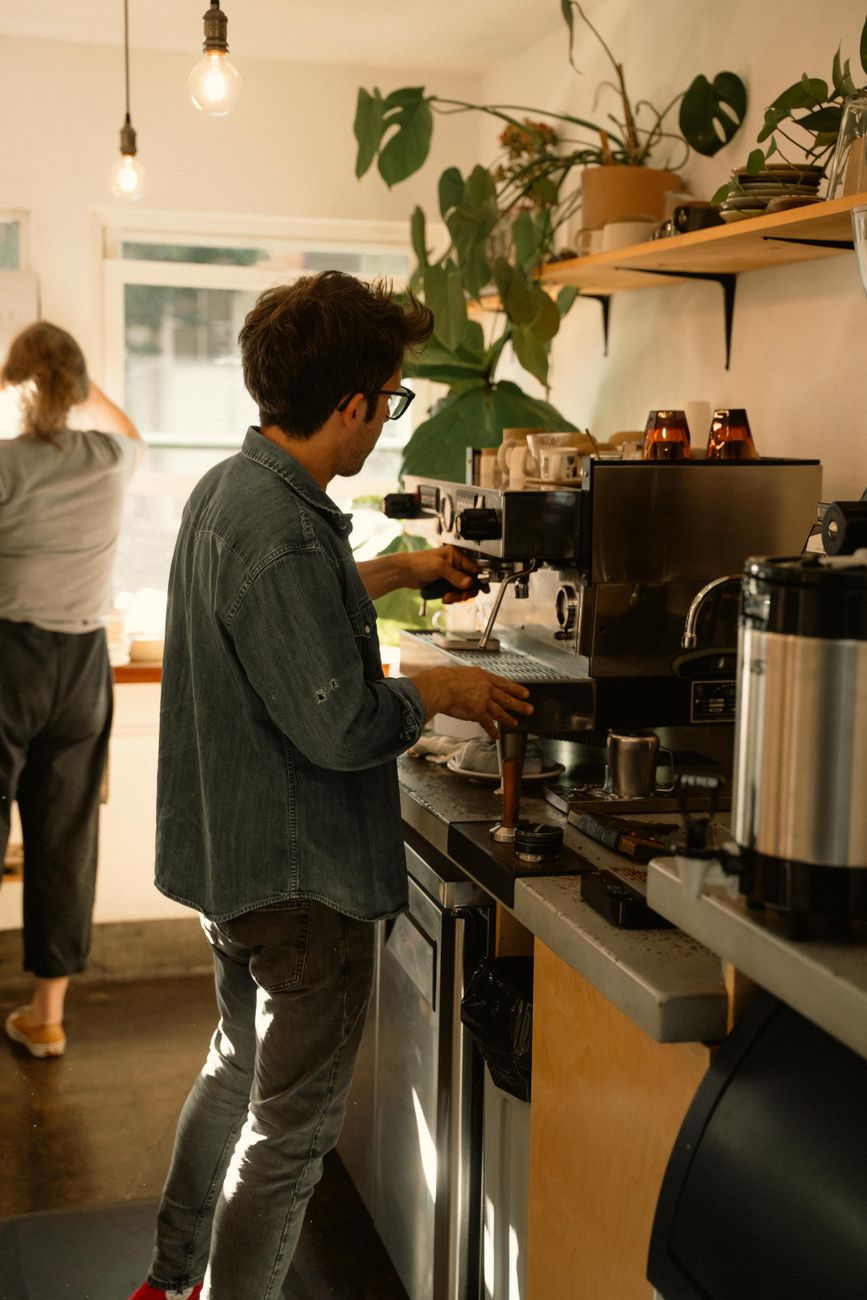 Barista pulling a shot at the espresso machine in a plant-filled sunlit corner