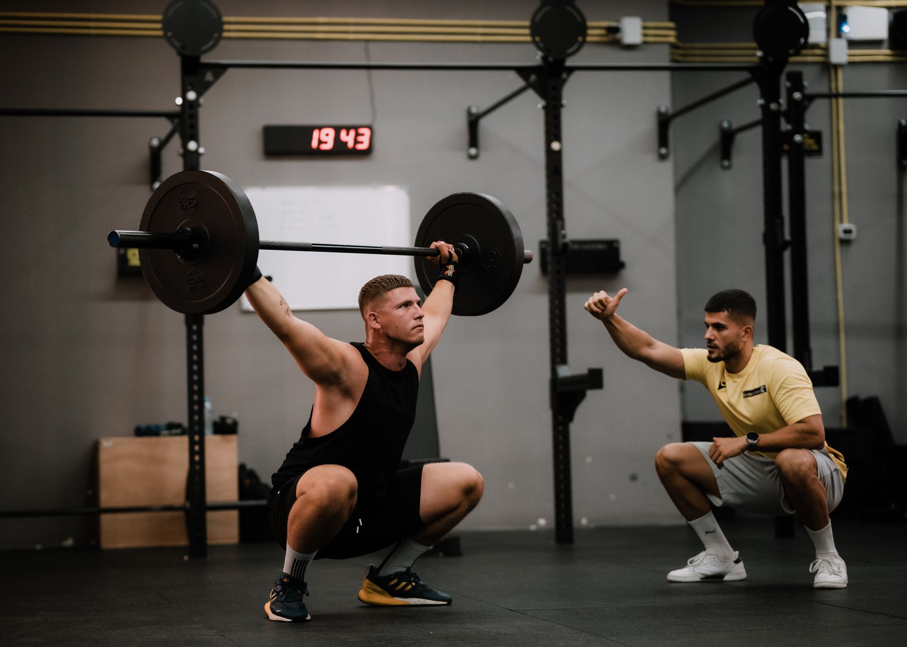 A coach crouches to cue an athlete holding a barbell overhead squat