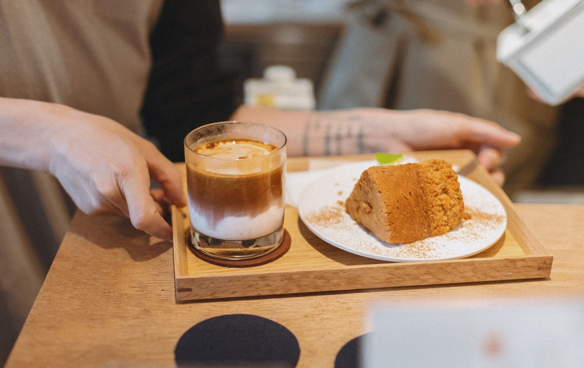 Hands passing a timber tray with an iced latte and a slice of cake