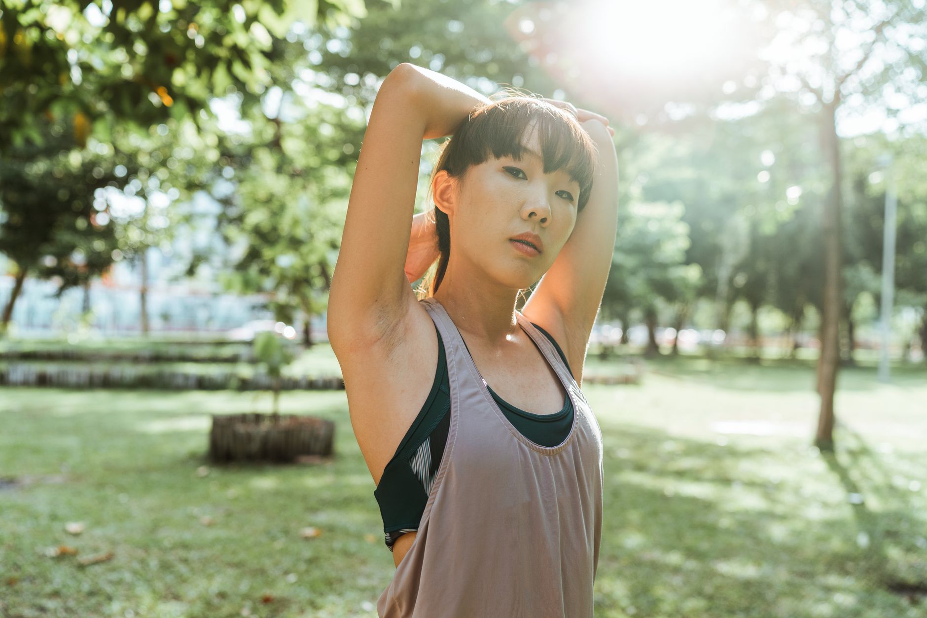 A woman stretches her arms overhead while warming up in a sunlit park