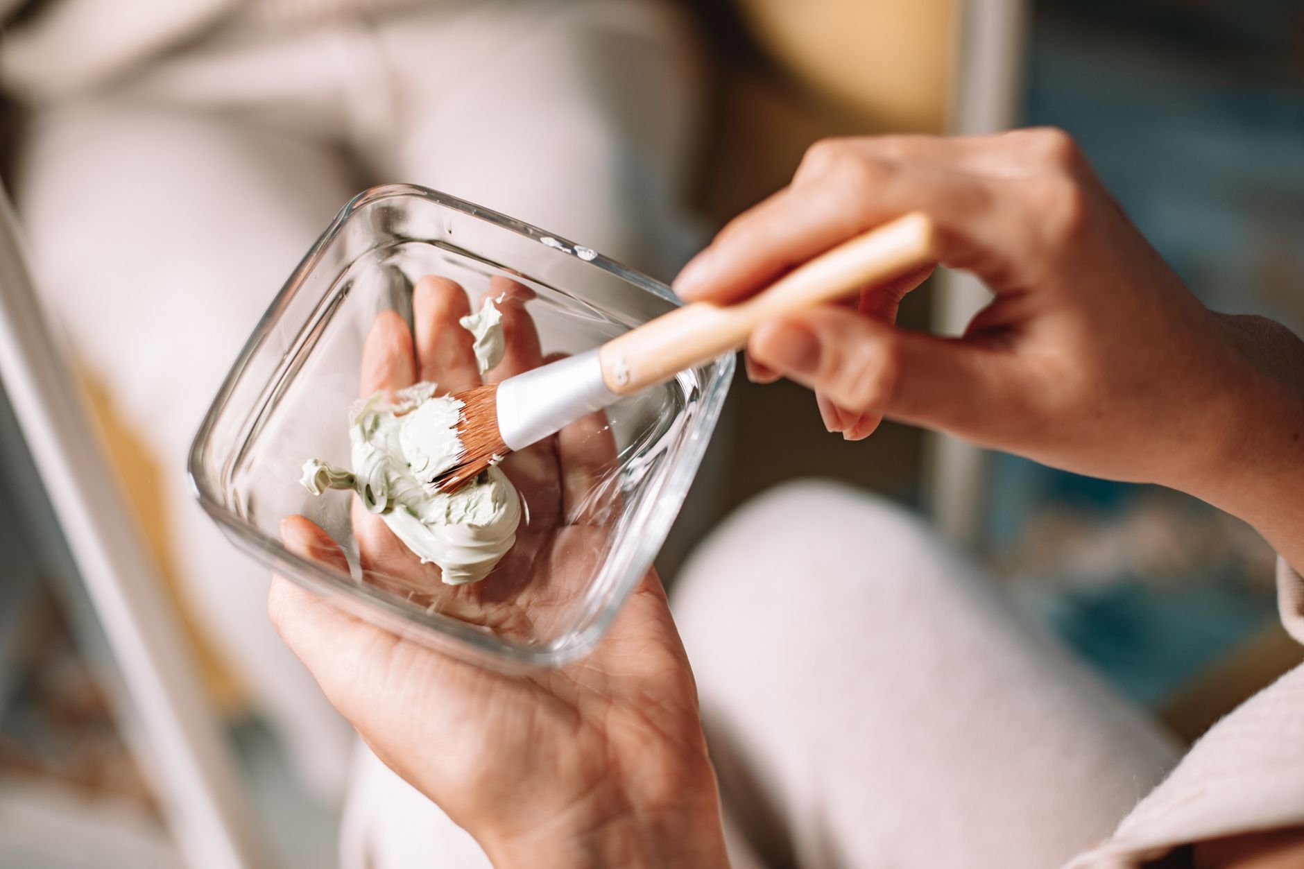 Hands mix a sage green botanical mask in a glass bowl with a small brush