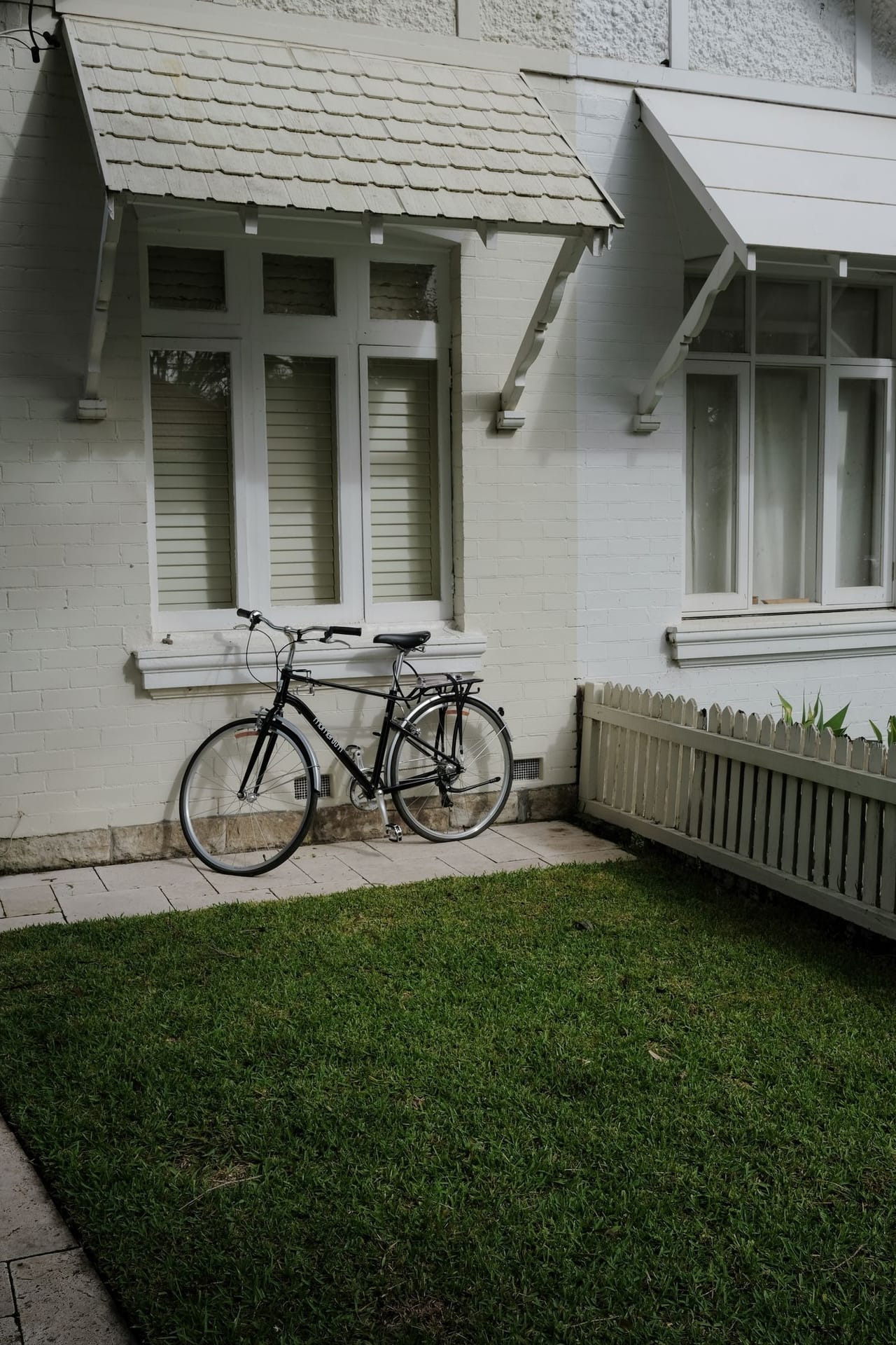 A white-painted period home with a tiled awning, timber windows and a picket fence beside a green lawn