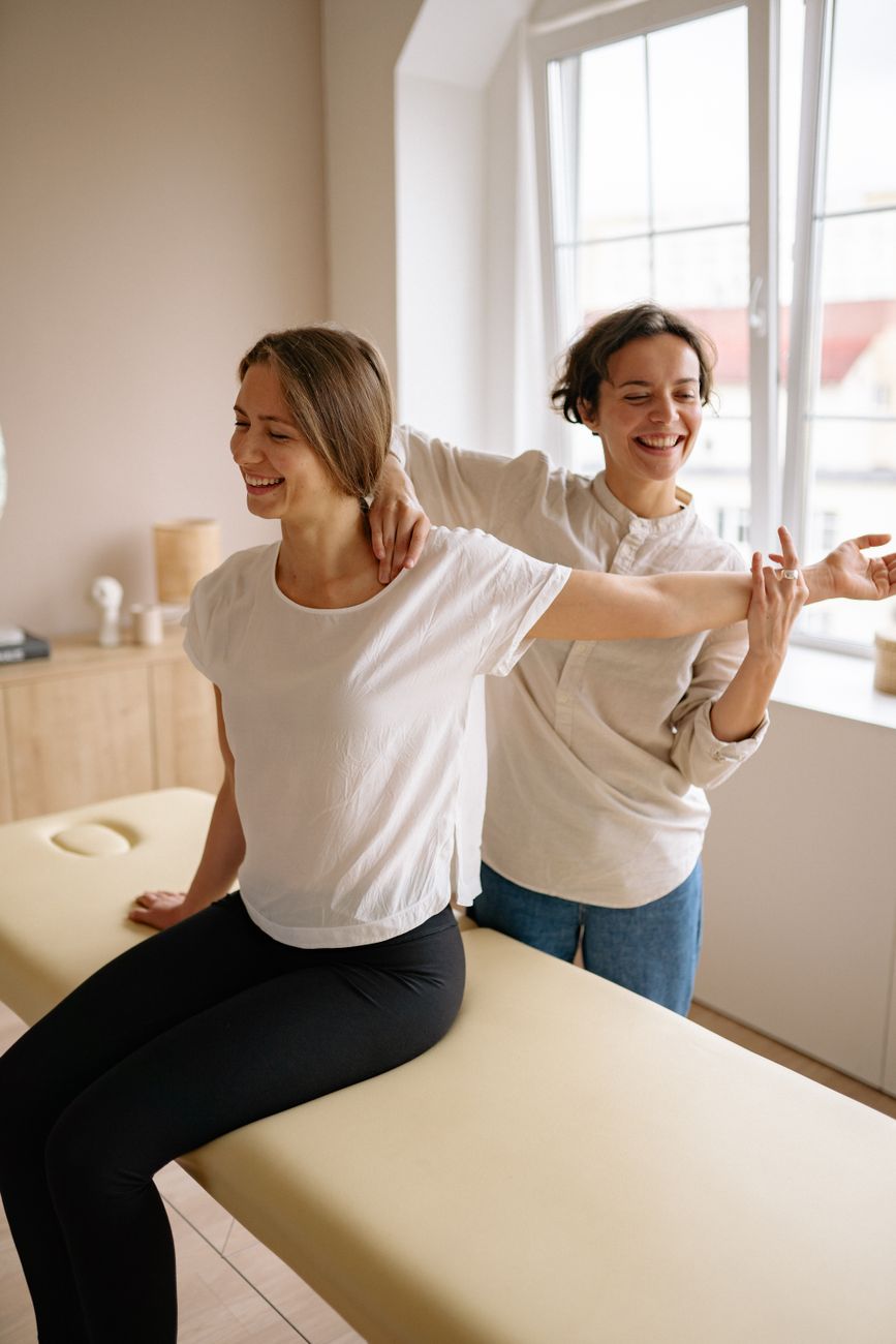 A physiotherapist guides a smiling patient through a shoulder stretch beside a window