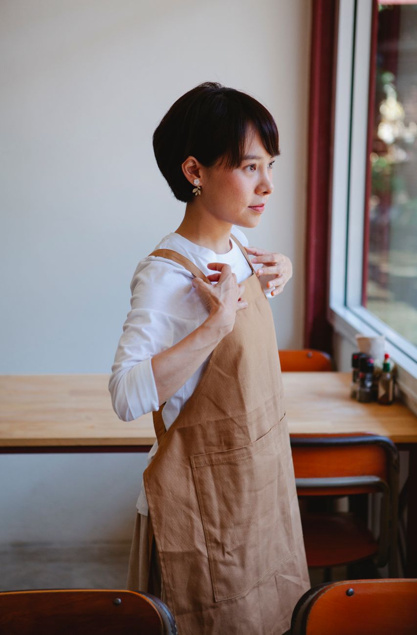 Cafe owner in a tan apron pausing by the front window in soft morning light