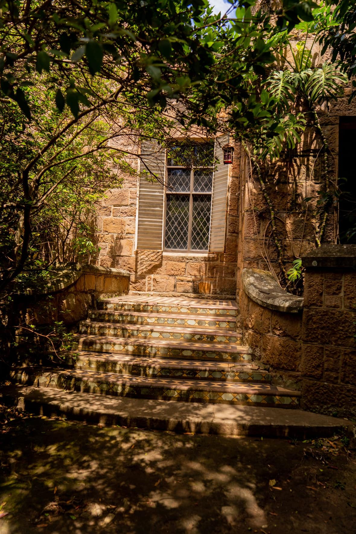 A sunlit sandstone home with tessellated tiled steps and a leadlight window framed by lush garden