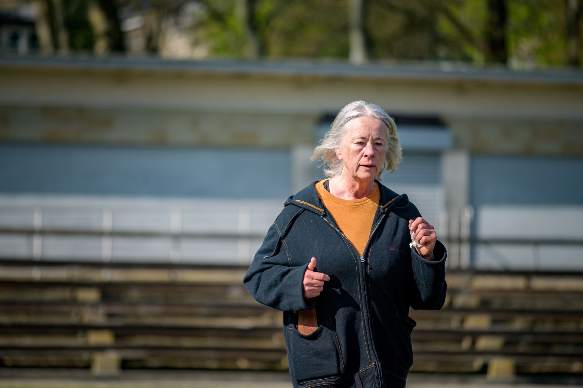 An older woman jogs steadily past a grandstand on a sunny morning