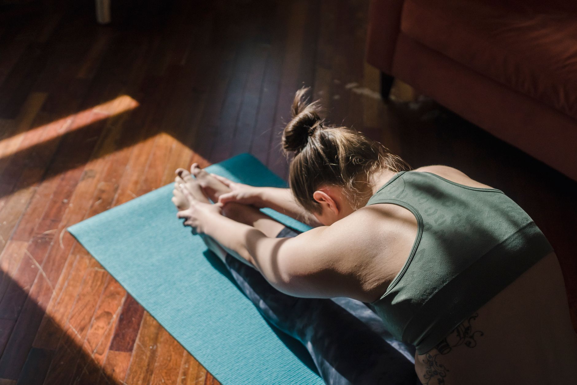 A woman folds forward over her legs on an exercise mat in a sunlit lounge room